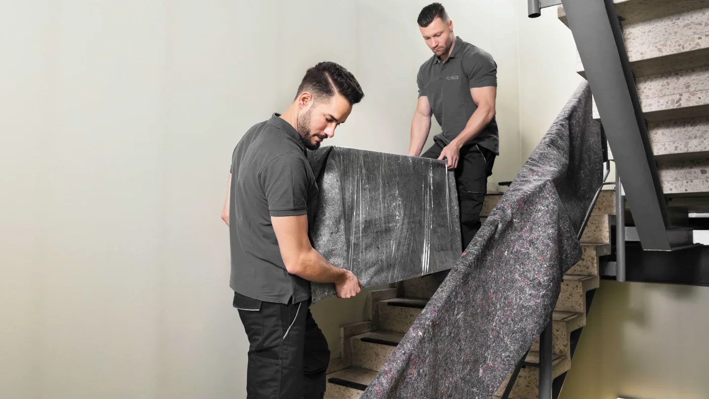 Elite Anywhere handler carrying blanket-wrapped furniture up a stairwell during a final-mile delivery